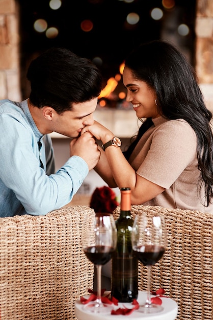 Pareja joven en una cita romántica en un restaurante por la noche y besándose en la mano en la chimenea del café con copas de vino Gente feliz enamorada día de san valentín juntos y novia sonríen al guapo novio