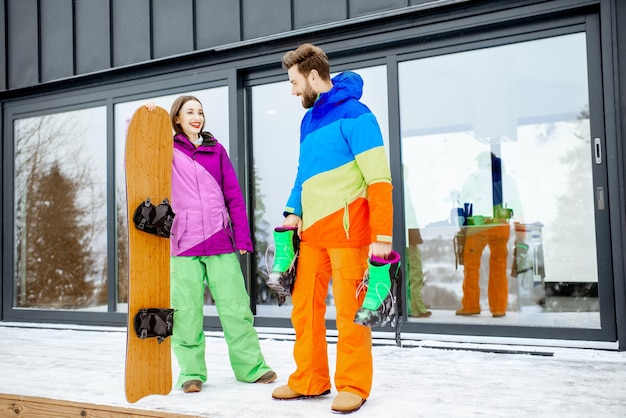 Pareja joven en coloridos trajes de esquí de pie junto con snowboard cerca de la moderna casa u hotel en las montañas