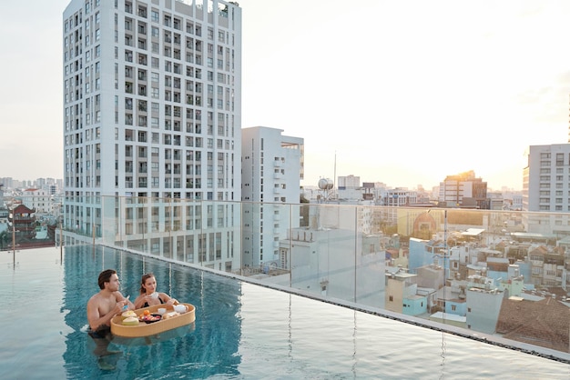 Pareja joven comiendo un delicioso desayuno cuando está de pie en la piscina de la azotea y disfruta ...