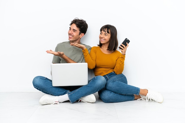 Pareja joven con una computadora portátil y un móvil sentados en el suelo apuntando hacia atrás y presentando un producto