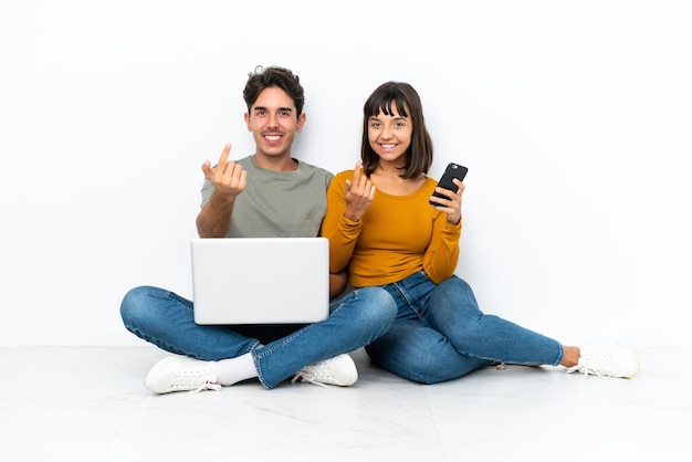 Pareja joven con una computadora portátil y un móvil sentados en el suelo invitando a venir con la mano Feliz de que hayas venido