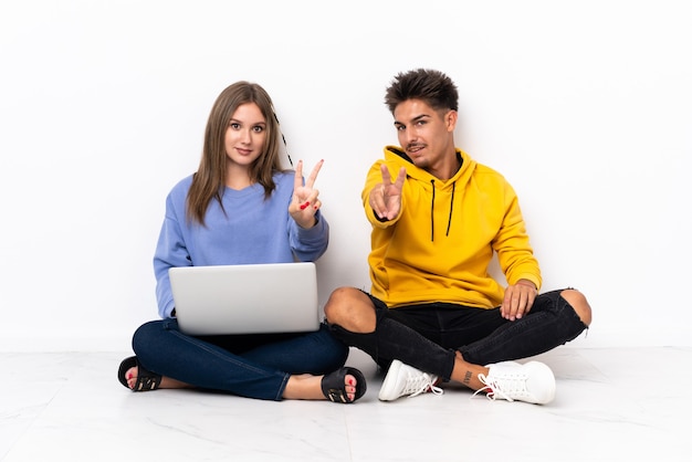 Pareja joven con una computadora portátil sentada en el suelo en blanco sonriendo y mostrando el signo de la victoria