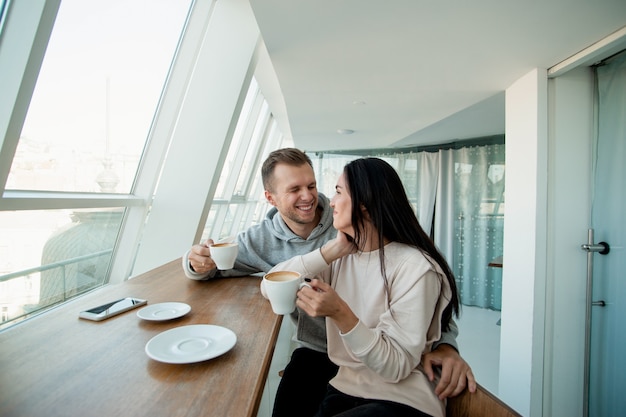 Pareja joven coqueteando en la cafetería. Hermosa mujer con cabello largo y oscuro y un hombre guapo con un elegante corte de pelo se miran, sostienen las tazas de café y sonríen con alegría. Concepto de citas.