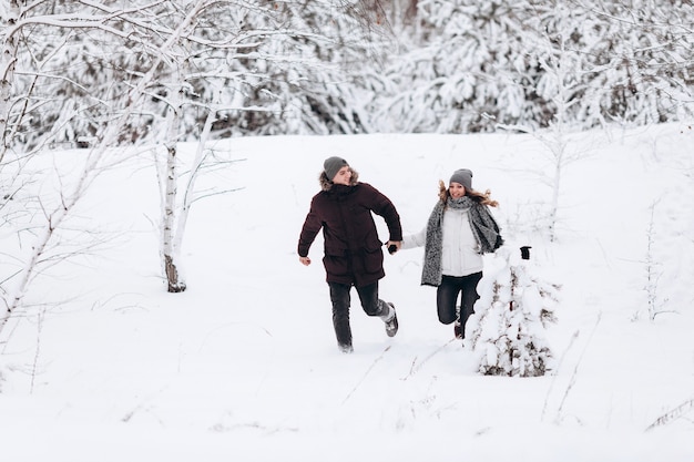 Pareja joven corriendo en un campo de invierno cubierto de nieve cerca de pino