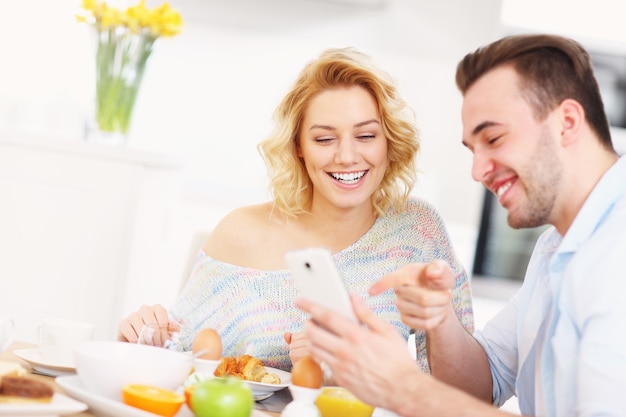 Pareja joven desayunando en la cocina y usando un teléfono inteligente.