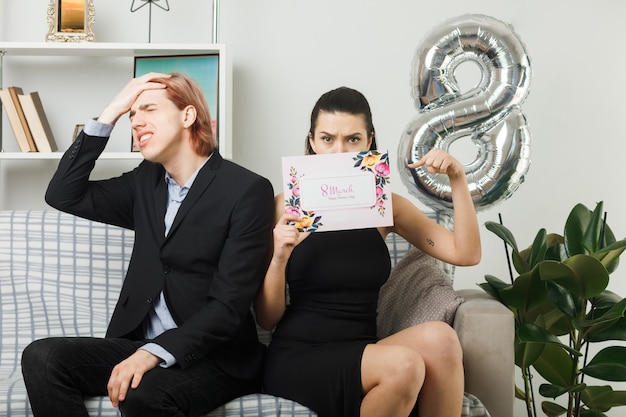 Pareja joven en el día de la mujer feliz celebración estricta y rostro cubierto con postal lamentado chico poniendo la mano en la frente sentado en el sofá en la sala de estar