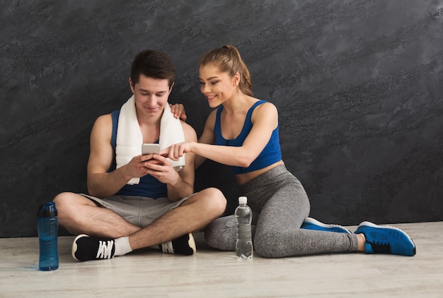 Pareja joven discutiendo el plan de entrenamiento, sentados en el suelo en el gimnasio. Hombre y mujer con smartphone, haciendo horarios para entrenamientos personales, espacio de copia