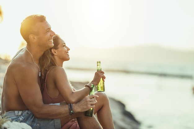 Pareja joven divirtiéndose y bebiendo cerveza en la playa junto al mar. Disfrutando de su amor.