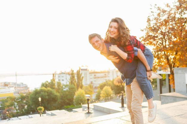 Pareja joven divirtiéndose en la ciudad al amanecer. Un hombre guapo dando a su novia un paseo a cuestas. Disfrutando cada momento juntos.