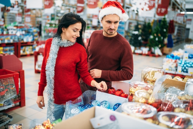 Pareja joven elige juguetes para árboles de Navidad en la tienda, tradición familiar. Compras de diciembre de artículos y decoraciones navideñas