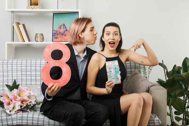 Pareja joven emocionada en el día de la mujer feliz sosteniendo el número ocho con presente sentado en el sofá en la sala de estar