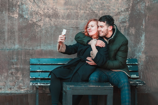 Pareja joven emocional alegre sentado en el banco y hacer Selfie. Historia de amor de dos personas felices en la calle