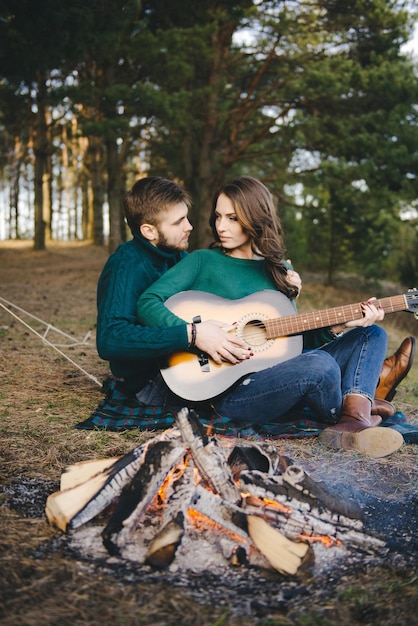 Pareja joven enamorada acampando turistas sentados junto a una fogata contra una carpa en el bosque tocando la guitarra