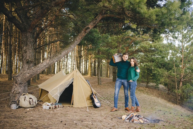 Pareja joven enamorada acampando turistas sentados junto a un fuego contra una carpa en el bosque escuchando radio retro