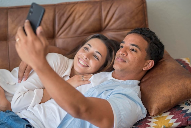 Pareja joven enamorada disfruta y usa el teléfono móvil moderno juntos acostados y relajados en el sofá Amor y relación jóvenes actividad de ocio interior con conexión a internet Concepto de videollamada