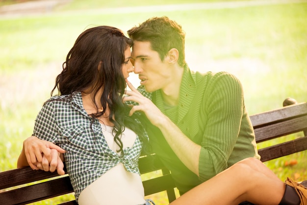 Una pareja joven enamorada sentada en un banco del parque, iluminada por la luz del sol, se mira apasionadamente el momento antes del beso.