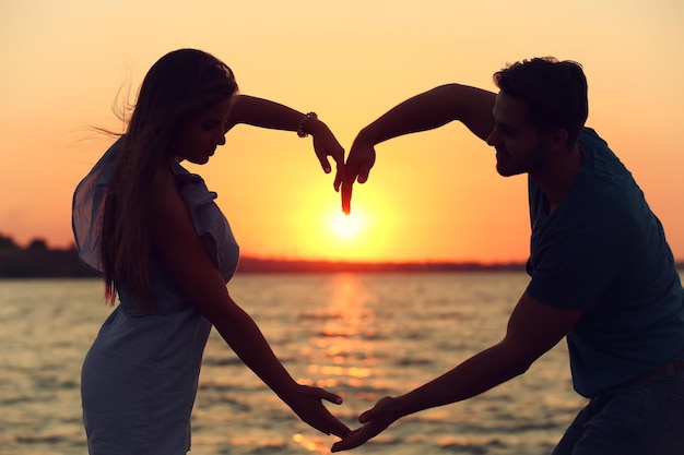 Una pareja joven enamorada tomándose de la mano en forma de un gran corazón en la playa al atardecer