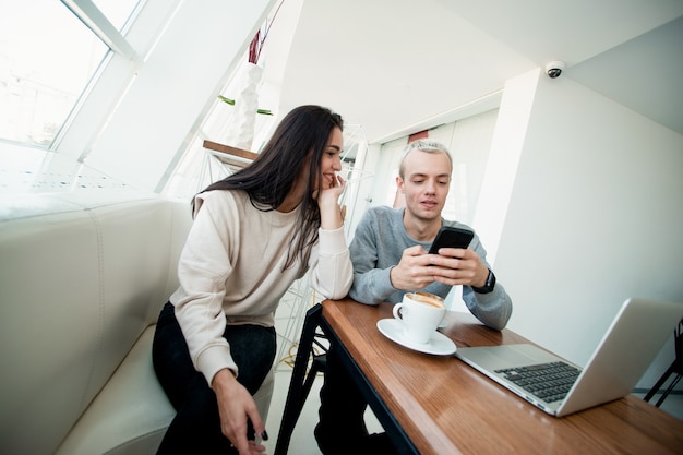 Foto pareja joven se encuentra en la cafetería. hombre rubio que muestra algo divertido en su teléfono inteligente a una mujer atractiva morena. portátil moderno en la mesa. concepto de redes sociales. mujer apoyada en su mano y sonríe.