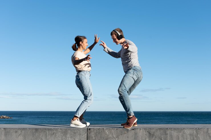 Una pareja joven escuchando música y bailando en un asiento de piedra a la orilla del mar en un día soleado