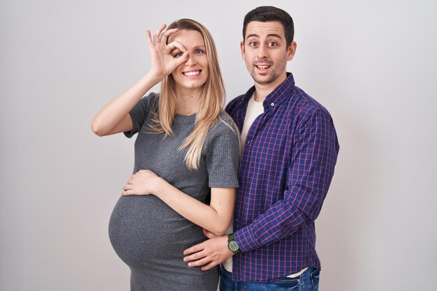 Una pareja joven esperando un bebé parado sobre fondo blanco haciendo un buen gesto con la mano sonriendo, mirando a los ojos a través de los dedos con cara feliz.