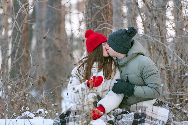 Una pareja joven está sentada abrazándose y besándose en un parque cubierto de nieve envolviéndose en un plaid Hombre y mujer en un picnic en el bosque de invierno