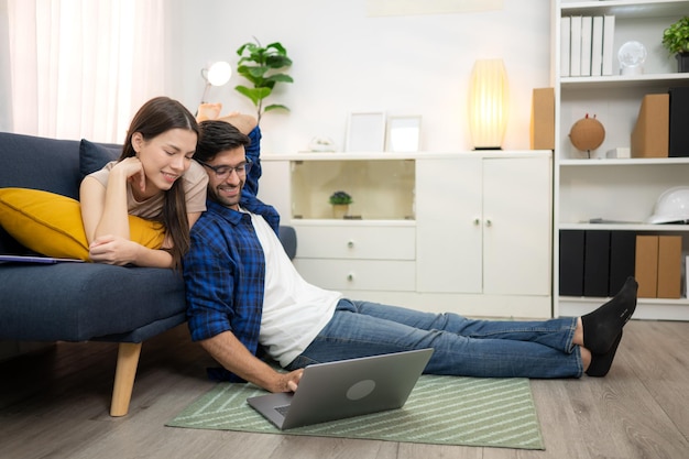 Una pareja joven está sonriendo mientras se relaja y trabaja en línea a través de una computadora portátil en la sala de estar de su casa
