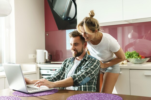 Pareja joven y feliz usando laptop en la cocina por la mañana