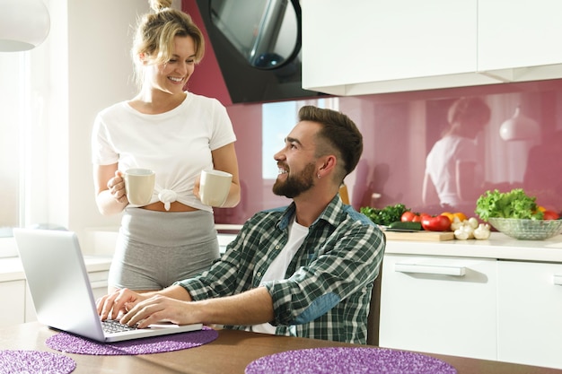 Pareja joven y feliz usando laptop en la cocina por la mañana