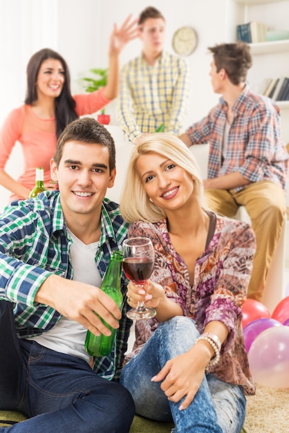 Pareja joven en la fiesta de casa, sentarse en el suelo, tostar con bebidas, sonriendo mirando a la cámara. Al fondo puedes ver a sus amigos que están sentados en el sofá.