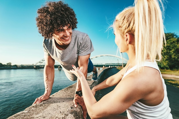 Una pareja joven de fitness está haciendo ejercicio en la pared junto al río en una puesta de sol. El hombre está agachado y sostiene pesas rusas, y la mujer lo sostiene.