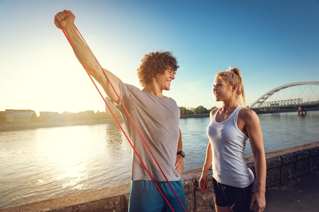 Una pareja joven de fitness haciendo ejercicio con una banda elástica junto al río al atardecer. El hombre estira los brazos y la mujer lo apoya.