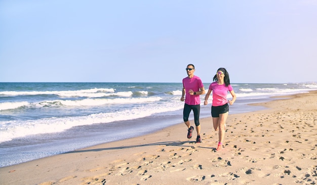 Pareja joven en forma corriendo en la playa durante el amanecer. Comienzo saludable del día. Vistiendo ropa deportiva rosa y negra. Corriendo por el mar.