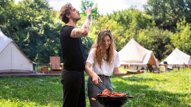 Pareja joven friendo carne a la parrilla y bebiendo cerveza. Verdor alrededor. Glamping