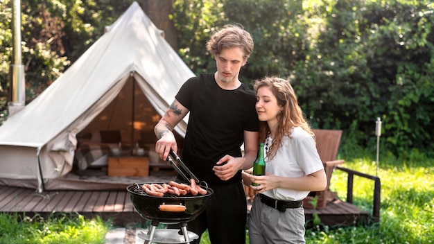 Pareja joven friendo carne a la parrilla y bebiendo cerveza. Verdor alrededor. Glamping