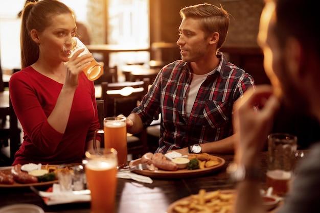Pareja joven hablando entre ellos mientras beben cerveza y almuerzan en una taberna con amigos