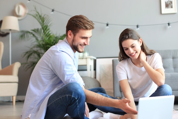 Pareja joven haciendo algunas compras en línea en casa, usando una computadora portátil en el piso.