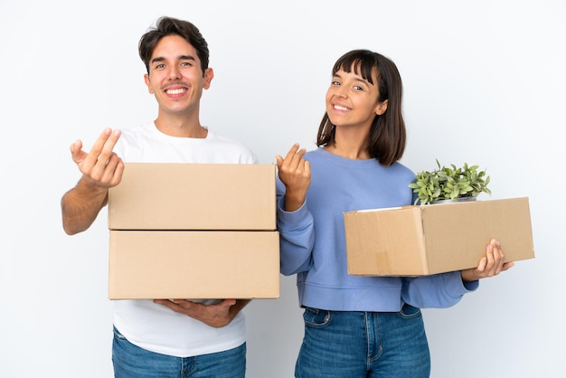 Pareja joven haciendo un movimiento mientras recoge una caja llena de cosas aisladas en fondo blanco invitando a venir con la mano Feliz de que hayas venido