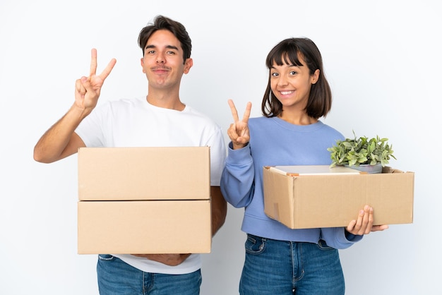 Pareja joven haciendo un movimiento mientras recoge una caja llena de cosas aisladas sobre fondo blanco sonriendo y mostrando el signo de la victoria