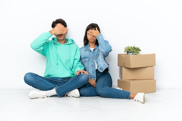 Pareja joven haciendo un movimiento mientras recoge una caja llena de cosas sentadas en el suelo aisladas en un fondo blanco cubriendo los ojos con las manos. no quiero ver algo