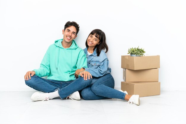 Pareja joven haciendo un movimiento mientras recoge una caja llena de cosas sentadas en el suelo aisladas en fondo blanco manteniendo los brazos cruzados en posición lateral mientras sonríe