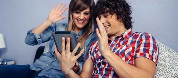 Pareja joven haciendo una videollamada con la tableta sentada en la cama