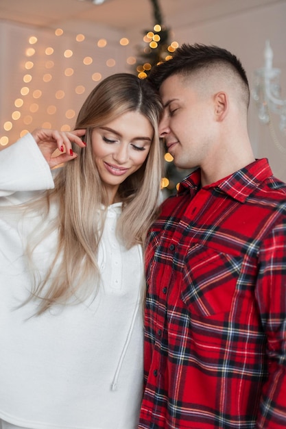 Una pareja joven y hermosa de moda con ropa elegante y cómoda se para y se abraza cerca del árbol de Navidad en casa