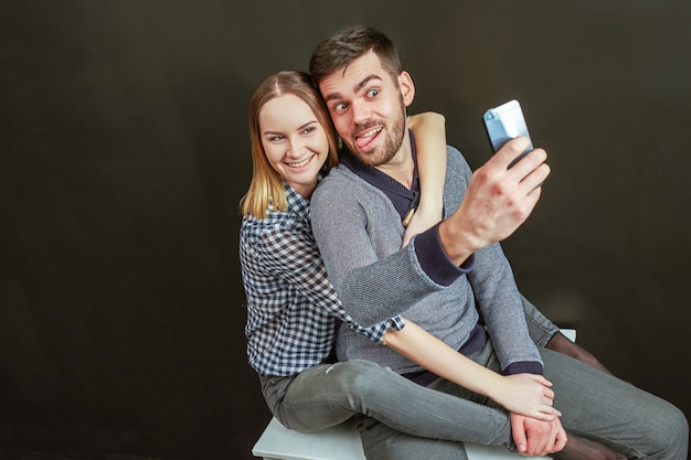 Pareja joven de hombre guapo con barba y mujer rubia sentada contra de fondo negro y haciendo un divertido selfie. Tiro del estudio