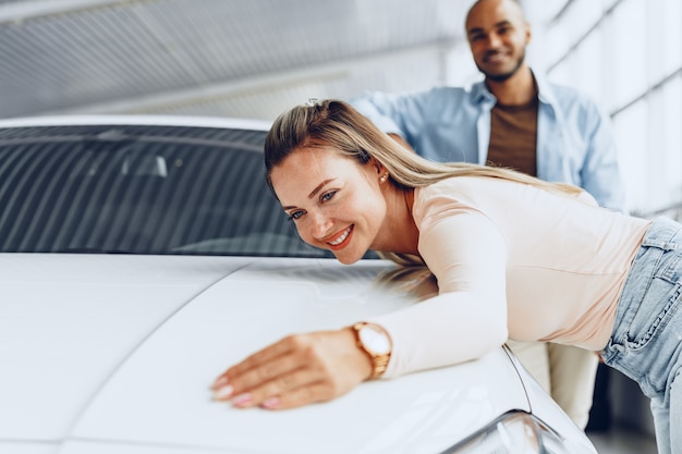 Pareja joven hombre y mujer abrazando su coche nuevo en una tienda de coches de cerca