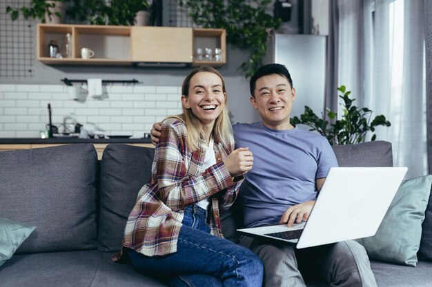 Pareja joven hombre y mujer asiáticos en casa sentados en un sofá juntos usando una laptop familia multirracial feliz y sonriente mirando la cámara