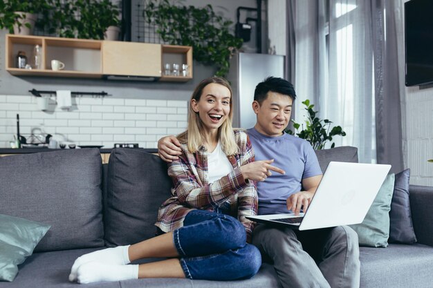 Pareja joven hombre y mujer asiáticos en casa sentados en un sofá juntos usando una laptop familia multirracial feliz y sonriente mirando la cámara