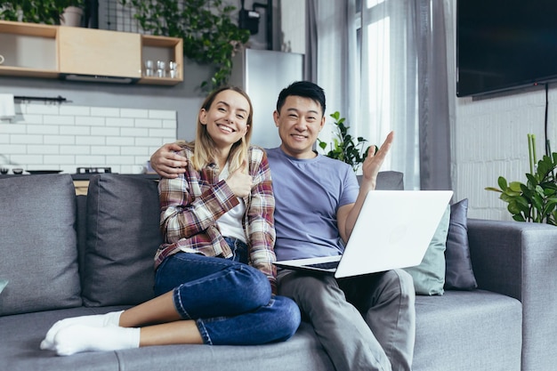 Pareja joven hombre y mujer asiáticos en casa sentados en un sofá juntos usando una laptop familia multirracial feliz y sonriente mirando la cámara