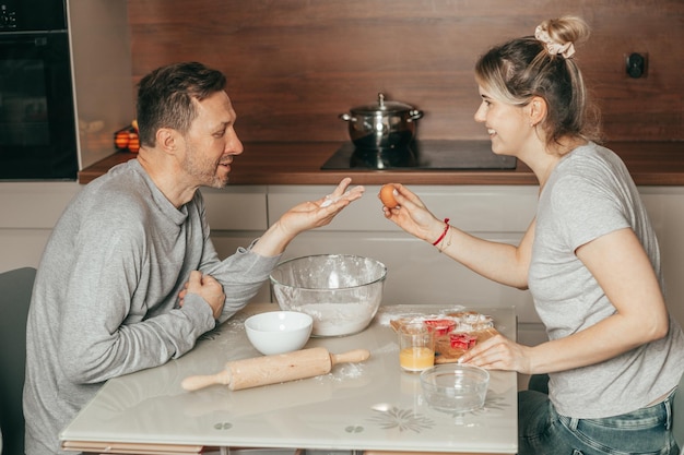 Pareja joven, hombre y mujer Diviértete hablando. Hornear en casa, mujer sosteniendo huevo en las manos, hornear galletas, cocinar en la cocina. Lindos hombres y mujeres trabajando juntos como equipo, gente feliz