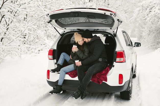 Una pareja joven, un hombre y una mujer, están sentados en el maletero de un coche en un bosque nevado de invierno