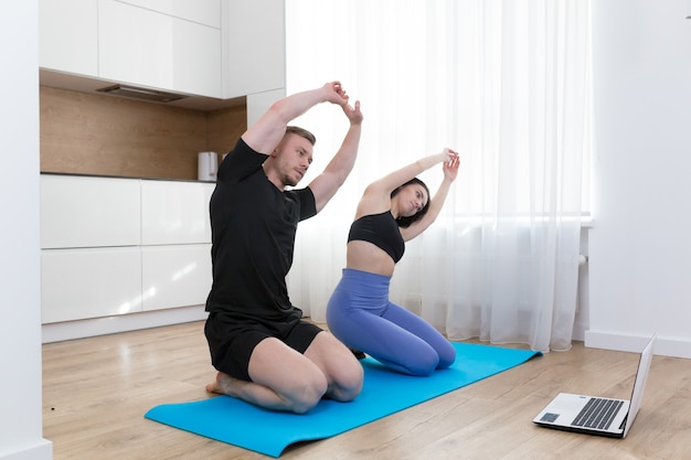 Pareja joven hombre y mujer haciendo fitness en casa en línea usando la computadora portátil, haciendo en línea con el entrenador en el piso en casa deportes